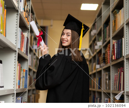 Happy young woman in graduate gown holding diploma in the library. Happy young woman in graduate gown holding diploma in the library. 102136094