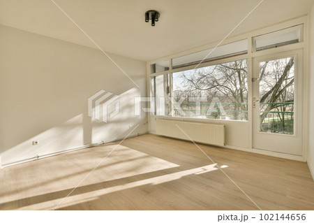 an empty living room with wood flooring and large windows looking out onto the trees in the distance are bare branches an empty living room with wood flooring and large windows looking out onto the trees in the distance are bare branches 102144656