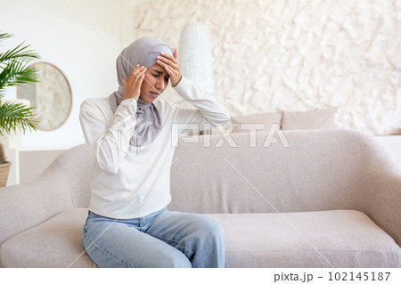 A young woman in a hijab is sitting on the sofa at home and holding her head with her hands, feeling severe pain, giving herself a massage, grimacing from tension. Stresses, worries, depresses. A young woman in a hijab is sitting on the sofa at home and holding her head with her hands, feeling severe pain, giving herself a massage, grimacing from tension. Stresses, worries, depresses. 102145187