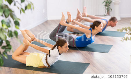 Diligent preteen girl practicing bow pose of yoga with her family Diligent preteen girl practicing bow pose of yoga with her family 102153795