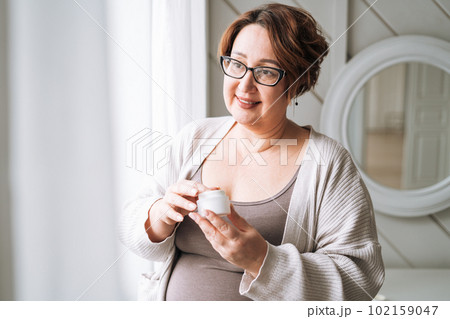 Smiling middle aged plus size woman with day cream near window at home Smiling middle aged plus size woman with day cream near window at home 102159047