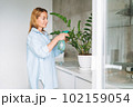 Young woman in blue shirt with spray with water in hands takes care of houseplant in kitchen at home 102159054