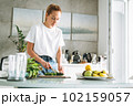 Young woman in white t-shirt and blue jeans cooking healthy food with cucumber in kitchen at home 102159057
