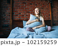 Young adult smiling woman with blonde long hair in casual sitting on bed with pillow at the home 102159259