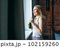 Young adult woman with blonde long hair in casual with green smoothie near window at the home 102159260