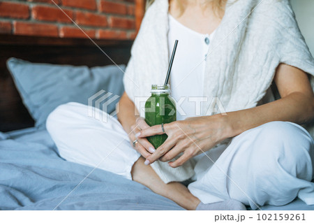 Crop photo of Young woman in casual clothes with green smoothie sitting on bed at the home 102159261