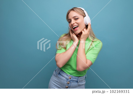 cute blond girl in casual outfit listens to music on the app in big white headphones on a blue cute blond girl in casual outfit listens to music on the app in big white headphones on a blue 102169331