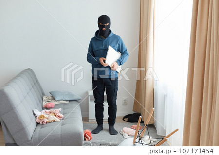 Man steals laptop in someone else house, runs away, being of persecution, wears robber mask and blue hoodie, standing near sofa and window, dangerous burglary breaking flat. Man steals laptop in someone else house, runs away, being of persecution, wears robber mask and blue hoodie, standing near sofa and window, dangerous burglary breaking flat. 102171454