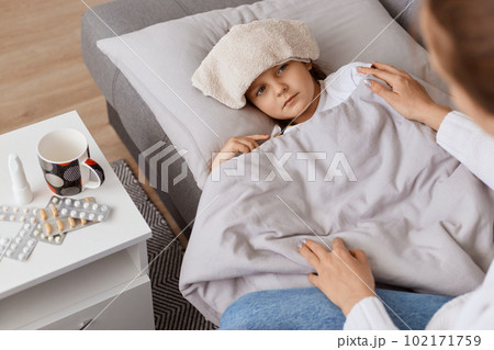 Indoor shot of unhealthy ill female child with wet towel on her forehead lying on sofa under blanket, mother sitting near her little sick girl, taking care and help. 102171759