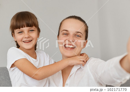 Smiling woman wearing white shirt having fun together with her daughter, broadcasting livestream or having video call, looking smiling at smartphone camera, POV. 102171770