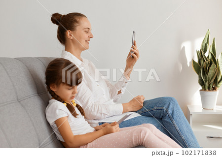 Indoor shot of beautiful smiling woman with bun hairstyle wearing white shirt and jeans sitting on sofa with her daughter, female having video call while kid choosing game to play. Indoor shot of beautiful smiling woman with bun hairstyle wearing white shirt and jeans sitting on sofa with her daughter, female having video call while kid choosing game to play. 102171838