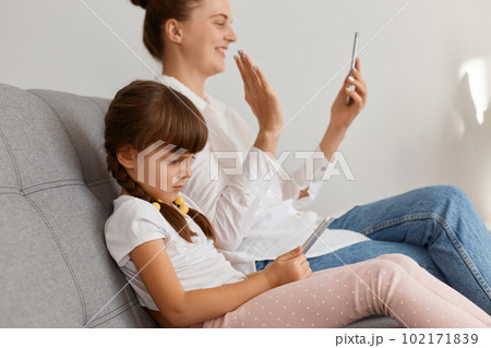 Side view portrait of positive attractive woman wearing white shirt and jeans sitting on sofa with her child, having video call or livestream, waving hand to followers. Side view portrait of positive attractive woman wearing white shirt and jeans sitting on sofa with her child, having video call or livestream, waving hand to followers. 102171839
