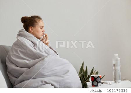 Side view portrait of sick upset woman with hair bun sitting on sofa wrapped in blanket using nasal spray, suffering runny nose and sneezing, catching cold. Side view portrait of sick upset woman with hair bun sitting on sofa wrapped in blanket using nasal spray, suffering runny nose and sneezing, catching cold. 102172352