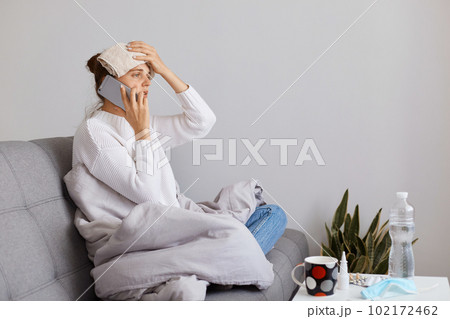 Unhealthy woman with bun hairstyle wearing white sweater sitting on cough, holding cell phone in hand and talking with doctor, keeping hand with towel on her forehead, having high temperature. 102172462