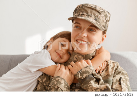 Indoor shot of happy positive military woman wearing camouflage uniform and cap, returning home from war or army, sitting on sofa with her kid, cute daughter misses mother and hugging her. Indoor shot of happy positive military woman wearing camouflage uniform and cap, returning home from war or army, sitting on sofa with her kid, cute daughter misses mother and hugging her. 102172618