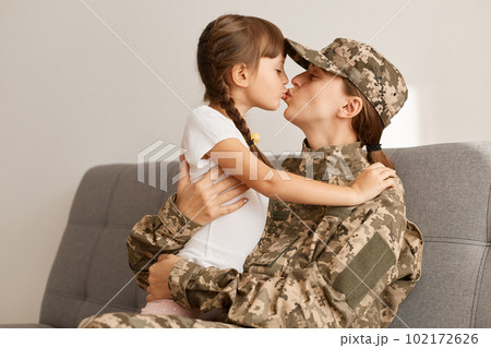 Indoor shot of happy military woman wearing camouflage uniform and cap, returning home from war or army, sitting on sofa with her daughter, kissing her child, missing, hugging each other. 102172626