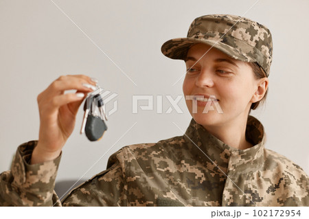 indoor shot of happy smiling woman soldier wearing camouflage uniform and hat, posing at home, holding and looking at key in hands, buying new flat or car. 102172954