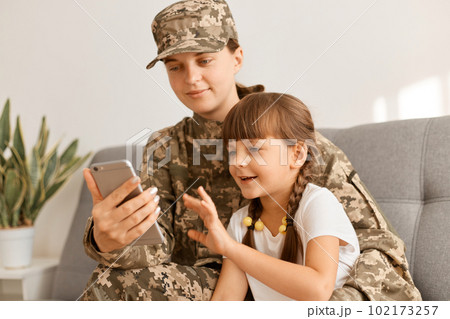 Portrait of young adult Caucasian military woman wearing camouflage uniform and cap posing with her daughter, mother holding mobile phone, kid trying to touch phone screen. 102173257