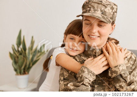 Horizontal shot of extremely happy satisfied woman wearing camouflage uniform and cap posing with her daughter, kid hugging her mother, military female arrived home from war or army. 102173267
