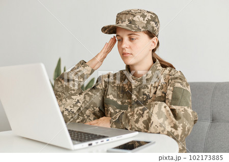 Horizontal shot of serious female soldier wearing military costume, posing indoor in light room, having online conversation and saluting to camera, saying yes sir. 102173885