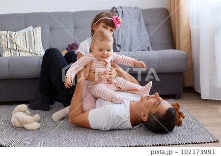 Extremely happy young adult man lying on floor near sofa and playing with his little kids, wearing white t shirt and trousers, children sitting on father, family enjoying spending time together. Extremely happy young adult man lying on floor near sofa and playing with his little kids, wearing white t shirt and trousers, children sitting on father, family enjoying spending time together. 102173991