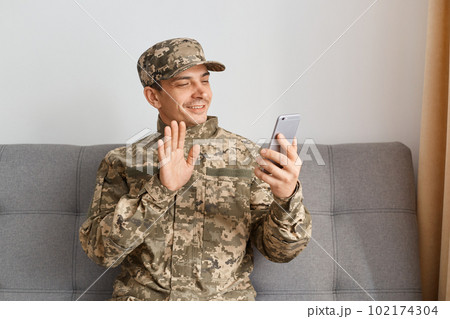 Portrait of smiling positive military man wearing camouflage uniform sitting on sofa, soldier talking on video call, saying hello or bye, looking at device screen with positive expression. Portrait of smiling positive military man wearing camouflage uniform sitting on sofa, soldier talking on video call, saying hello or bye, looking at device screen with positive expression. 102174304