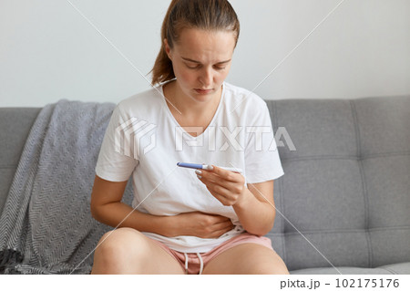 Sad unhappy woman wearing while casual style t shirt waiting for pregnancy test result thinking about future, touching her belly while sitting on sofa in living room alone. Sad unhappy woman wearing while casual style t shirt waiting for pregnancy test result thinking about future, touching her belly while sitting on sofa in living room alone. 102175176