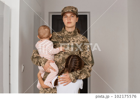 Serious proud military mother wearing camouflage uniform and hat, returning home after served in army, hugging her children after long-term separation. 102175576