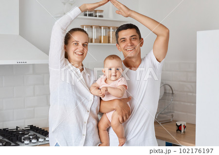 Indoor shot of happy smiling couple standing with baby daughter and making roof with hands above their heads, feels in safety together at home, expressing positive emotions. Indoor shot of happy smiling couple standing with baby daughter and making roof with hands above their heads, feels in safety together at home, expressing positive emotions. 102176197