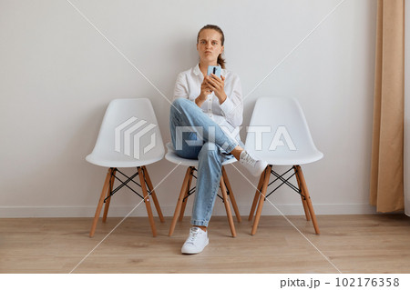 Confused sad attractive female with ponytail sitting on chair against light wall, having funny facial expression, woman wearing stylish jeans and white shirt, looking at camera. Confused sad attractive female with ponytail sitting on chair against light wall, having funny facial expression, woman wearing stylish jeans and white shirt, looking at camera. 102176358