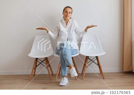 Indoor shot of confused helpless attractive female with ponytail sitting on chair with spreading hands against light wall, woman wearing stylish jeans and white shirt. Indoor shot of confused helpless attractive female with ponytail sitting on chair with spreading hands against light wall, woman wearing stylish jeans and white shirt. 102176365