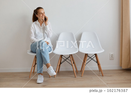 Portrait of adorable caucasian woman wearing white shirt and jeans sitting on chair in queue and talking phone, having conversation, looking away with smile, expressing positive emotions. 102176387