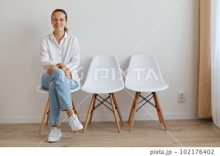 Portrait of happy smiling woman wearing white shirt and jeans sitting on chair in queue, holding her hands on her legs, looking at camera with optimistic expression. 102176402