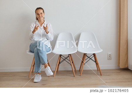 Full length portrait of satisfied young adult woman wearing white shirt and jeans sitting on chair in queue, looking and pointing at camera, being happy and delighted. 102176405