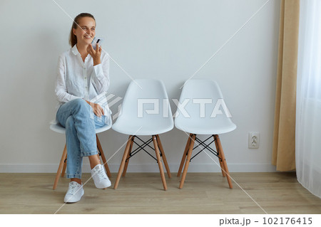 Portrait of smiling young adult woman sitting on chair, holding smart phone, asking voice assistant on cell phone, giving task, recording message, expressing positive emotions. Portrait of smiling young adult woman sitting on chair, holding smart phone, asking voice assistant on cell phone, giving task, recording message, expressing positive emotions. 102176415
