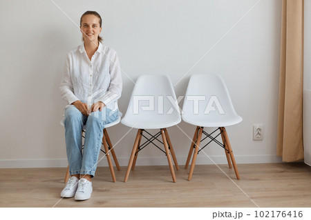 Indoor shot of calm Caucasian woman wearing white shirt and jeans sitting on chair in queue, looking at camera with smile and happy facial expression. Indoor shot of calm Caucasian woman wearing white shirt and jeans sitting on chair in queue, looking at camera with smile and happy facial expression. 102176416