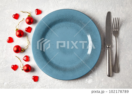 Empty blue ceramic plate with cutlery surrounded by red christmas balls and hearts on a light concrete table 102178789
