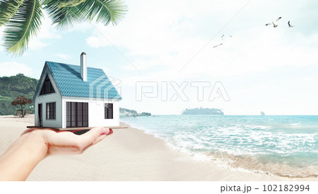 Moving to island. Female hand holding 3D model of small comfortable house against beach, palms and ocean background Moving to island. Female hand holding 3D model of small comfortable house against beach, palms and ocean background 102182994