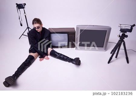 Young girl in stylish black jacket, leather boots and sunglasses sitting on floor around retro TV sets and camera 102183236