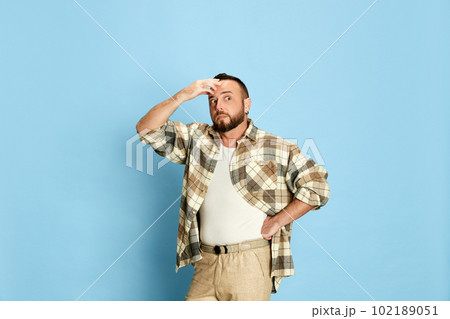 Portrait of bearded man in casual clothes holding hand to forehead, showing shocked emotions against blue studio background Portrait of bearded man in casual clothes holding hand to forehead, showing shocked emotions against blue studio background 102189051