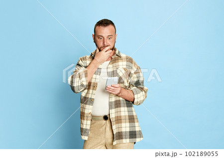 Portrait of bearded man in checkered shirt looking with shocked face on messages on mobile phone against blue studio background Portrait of bearded man in checkered shirt looking with shocked face on messages on mobile phone against blue studio background 102189055