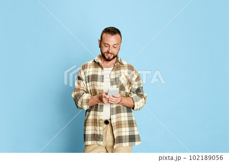 Portrait of happy, smiling, bearded man in checkered shirt typing messages with mobile phone against blue studio background Portrait of happy, smiling, bearded man in checkered shirt typing messages with mobile phone against blue studio background 102189056