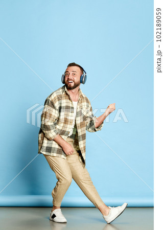 Portrait of bearded man in white t-shirt and checkered shirt listening to music in headphones against blue studio background Portrait of bearded man in white t-shirt and checkered shirt listening to music in headphones against blue studio background 102189059