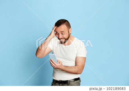 Portrait of happy, excited, bearded man in white t-shirt emotionally looking on mobile phone with shocked, positive face against blue studio background 102189083