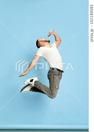 Full-length portrait of bearded energetic man in casual white t-shirt singing in microphone, jumping against blue studio background Full-length portrait of bearded energetic man in casual white t-shirt singing in microphone, jumping against blue studio background 102189085