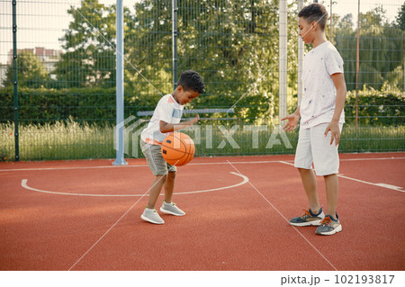 Two multiracional brothers playing basketball on a court near the park. Boys wearing white t-shirts. Older brother teach little one how to play basketball. 102193817