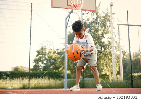Young boy standing on basketball court near the park. Boy wearing white t-shirt. Boy playing with basketball ball. Young boy standing on basketball court near the park. Boy wearing white t-shirt. Boy playing with basketball ball. 102193832
