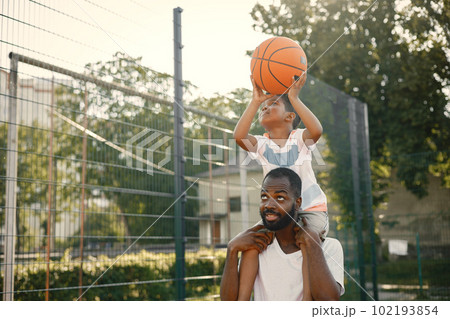 Black father holding on shoulders his little son to help him to score a basket. They standing on a basketball court. Man and boy wearing white t-shirts. 102193854