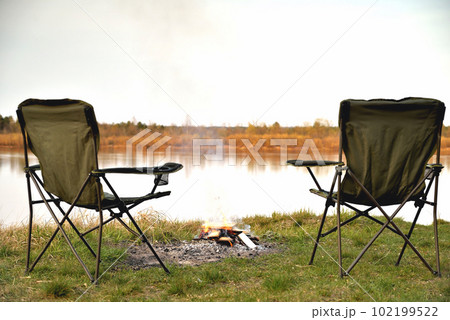 A pair of tourist armchairs, a folding chair, a bonfire on the river bank in the evening at sunset, twilight in autumn at sunset, relaxation and rest, the beginning of autumn, tourism and travel, a A pair of tourist armchairs, a folding chair, a bonfire on the river bank in the evening at sunset, twilight in autumn at sunset, relaxation and rest, the beginning of autumn, tourism and travel, a 102199522