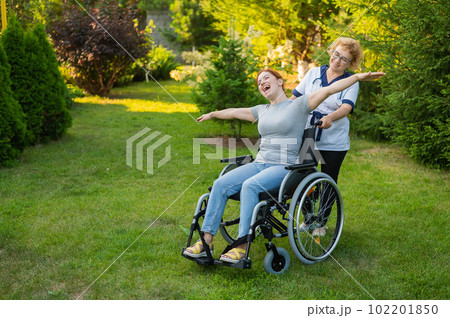 An elderly nurse walks with a middle-aged woman in a wheelchair through the park. The girl spread her arms outstretched like wings. An elderly nurse walks with a middle-aged woman in a wheelchair through the park. The girl spread her arms outstretched like wings. 102201850